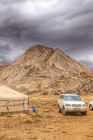 Three vehicles parked beside a yurt in Bayan-Unjuul, Töv, Mongolia, with mountains looming under a cloudy sky