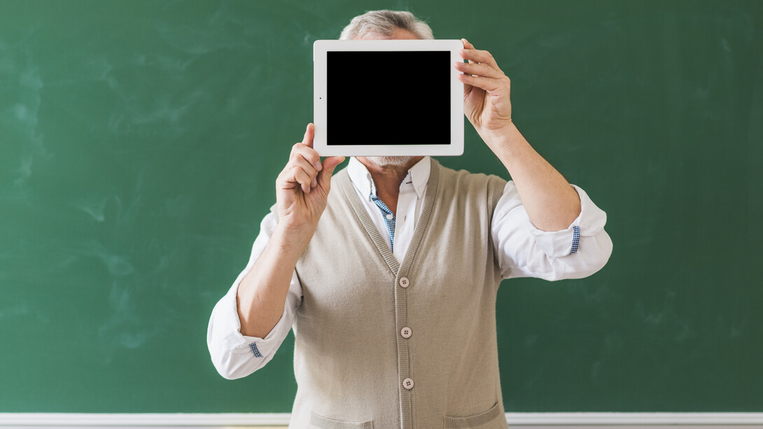 Un professore tiene un tablet con lo schermo nero nascondendo il viso, sullo sfondo una grande lavagna verde 