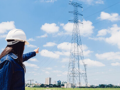 A woman wearing a safety helmet takes notes while inspecting a high-voltage power line, focusing on energy efficiency amid the ongoing energy crisis