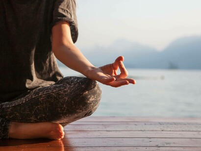 Dettaglio ravvicinato di una mano in Gyan Mudra (pollice e indice uniti) di una persona seduta con le gambe incrociate su una piattaforma di legno