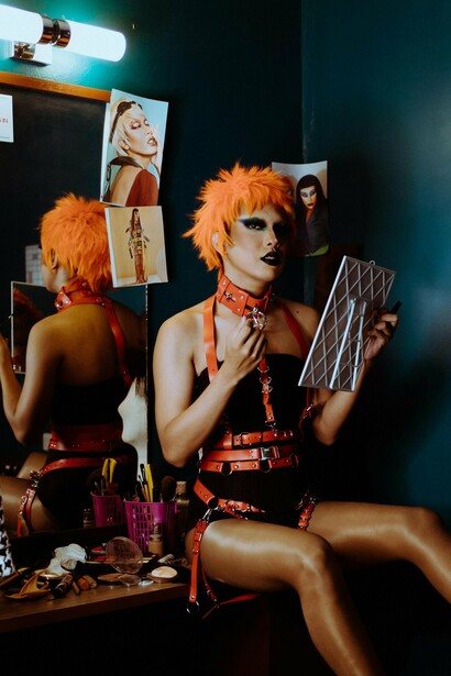 Surrounded by creative tools and inspirational imagery wearing an edgy orange wig, bold makeup, and a red harness ensemble, this striking individual exudes confidence and artistry and highlights the queer community’s commitment to pushing the boundaries of beauty and celebrating individuality through fearless creativity and style