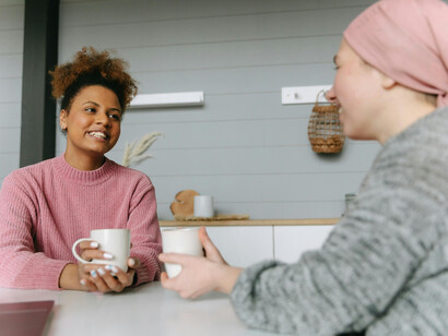 A woman in pink has a hot drink with a female cancer patient, both are sitting at a table