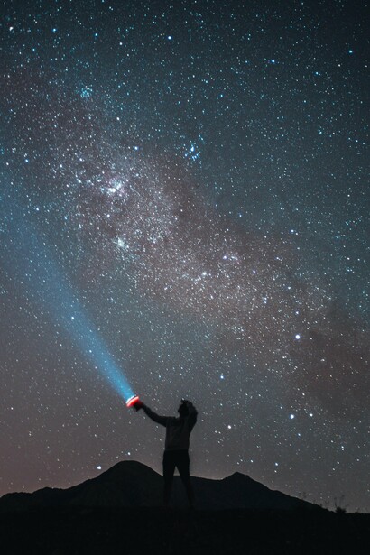 A figura solitária observa o céu estrelado enquanto projeta um feixe de luz na noite. Entre angústia e transcendência, o infinito continua a convocar o olhar humano