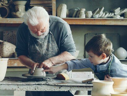 An older man guiding a young boy as he shapes clay on a pottery wheel, representing the passing of knowledge and craftsmanship between generations