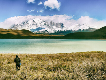 A person stands on lush green grass, overlooking a serene emerald lake with majestic mountain peaks in the background at Torres del Paine National Park, Chile, on a tranquil daytime scene