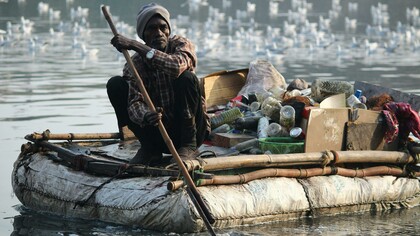 An adult man rows a makeshift raft filled with collected waste along a river, drawing attention to environmental pollution and cleanup efforts