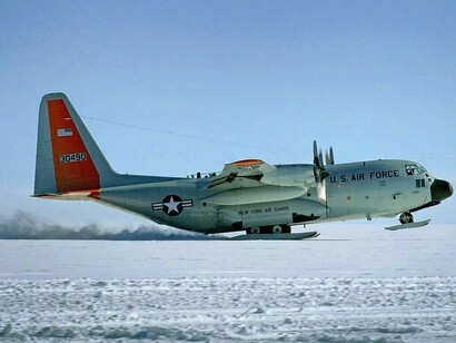 Um LC-130H Hercules decola da superfície gelada da Groenlândia, deixando um rastro de neve atrás de si. A aeronave simboliza a capacidade de alcançar e manter operações em áreas remotas do Ártico