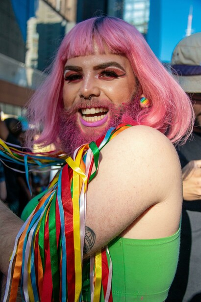 A smiling individual with bright pink hair, beard, and make-up during a pride parade captures the joyous spirit of queer identity, where style becomes a powerful medium for solidarity, celebration, and pushing boundaries in both personal and collective narratives