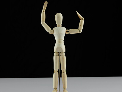 A wooden mannequin stands on a white table, its strings suggesting an unseen force controlling the crowd beyond