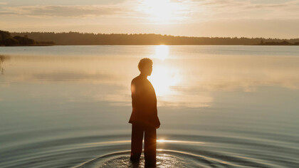 A figure stands at the shore of a lake, illuminated by the sun. 