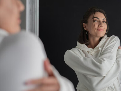 A young woman wearing a white hoodie, looking into a mirror as she explores self-discovery and self-reflection