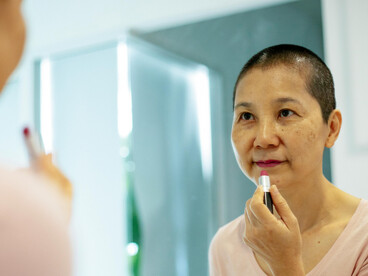 An Asian woman undergoing breast cancer treatment stands near a mirror, applying lipstick