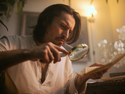 A thoughtful man scrutinizes a document with a magnifying glass under soft, warm lighting, surrounded by the timeless atmosphere of a classic study, reflecting careful analysis and intellectual curiosity