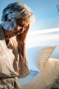 An elderly woman sits alone in a hospital room, holding her head in her hand
