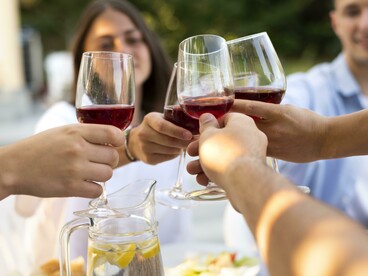 Gruppo di amici che brindano con calici di vino risso: "Il vino, la moda, il gusto e il tempo si fondono in una narrazione complessa e affascinante. Sono espressioni diverse dello stesso bisogno umano: quello di raccontarsi, di appartenere, di vivere con intensità e consapevolezza"
