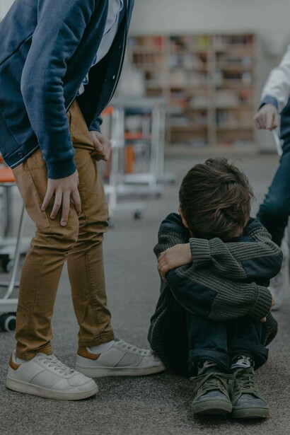 An older boy stands over a young kid huddled on the ground in fear, as he taunts and bullies hum