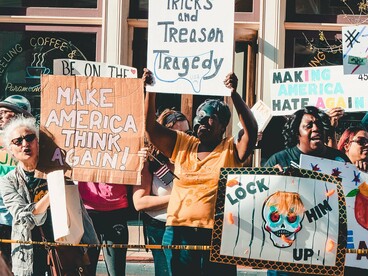 A diverse crowd raising hand-painted anti-Trump signs in public protest, representing growing resistance to political division and the erosion of democratic trust