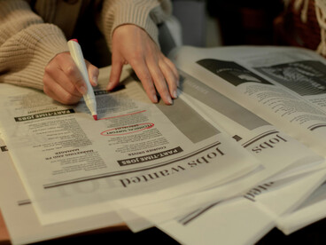 A close-up of a woman’s hand searching for job postings in a newspaper, reflecting the uncertainty of unemployment after graduation
