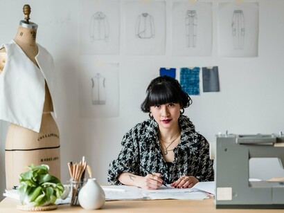 A fashion designer drawing a new garment while surrounded by sketches and materials, symbolizing the artistic intuition and technical knowledge that AI still depends on