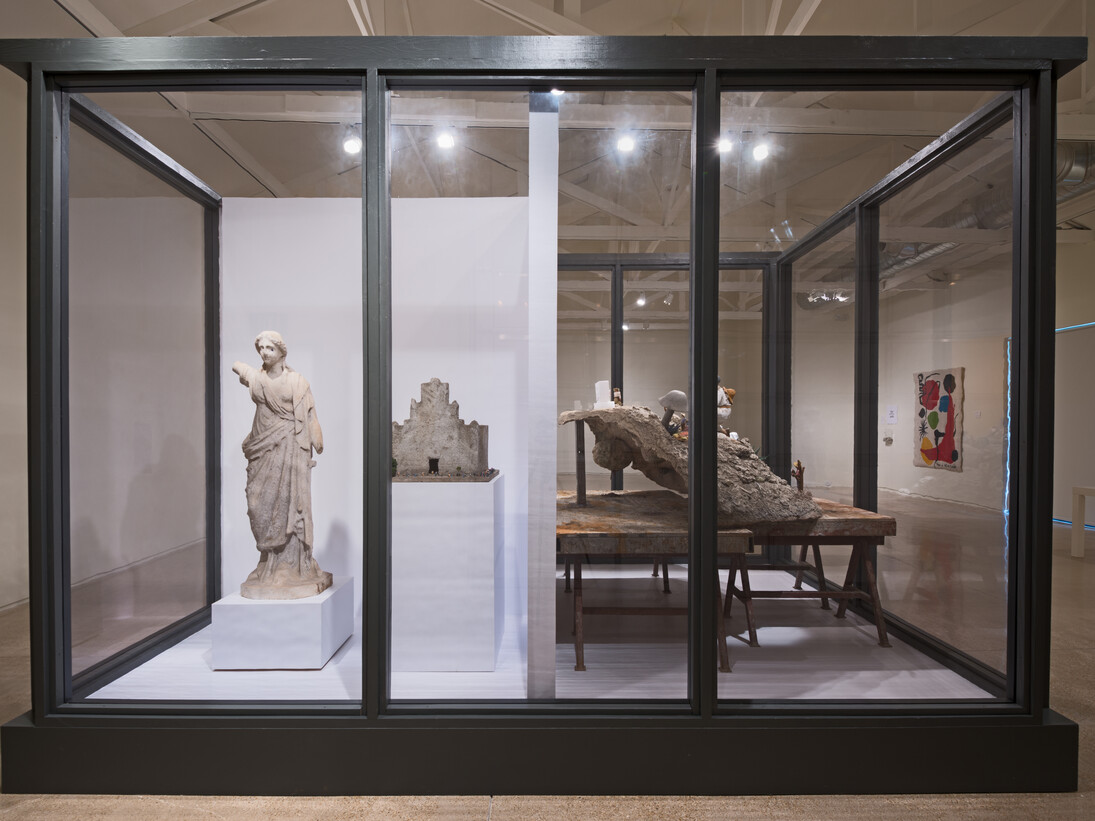 It Takes a Village, (2015), is a new, site-specific work installed in a large glass vitrine and includes various found objects, including Mexican clay figurines, artificial aloes, a plastic radio, a marble statue, Spanish moss, and Mexican colonial painting, among others