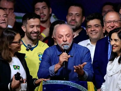 Lula discursa ao microfone, com uma das mãos estendida em um gesto de ênfase, enquanto aliados permanecem ao seu redor. À frente, uma bandeira brasileira cobre a parte superior da tribuna, trazendo à vista a inscrição “Ordem e Progresso”. A fotografia registra um momento de comunicação política em que a celebração da vitória se combina com a necessidade de apresentar os próximos passos. No horizonte de reflexão de Boaventura de Sousa Santos, a democracia não se esgota na escolha de representantes: depende também da capacidade de responder às desigualdades, ampliar a participação cidadã e submeter o exercício do poder ao controle público. A conquista eleitoral é, assim, um ponto de partida, não uma solução automática para os problemas do país