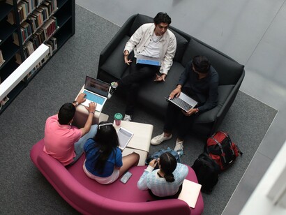 Students gathered around laptops and books in a shared learning space, representing knowledge as a collective resource for society
