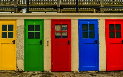 Uma sequência de portas de diferentes cores se alinha lado a lado em meio à paisagem urbana, criando uma composição marcada pela diversidade. Cada porta pode ser apenas uma escolha estética, mas também pode dizer algo sobre quem está do outro lado — afinal, como pergunta a autora, qual é a cor da porta da sua casa?