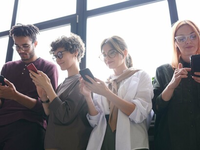 Quatro pessoas permanecem próximas, mas todas mantêm os olhos voltados para os smartphones que seguram nas mãos. A tecnologia ocupa o centro da atenção e transforma um momento compartilhado em uma experiência individual, marcada pela presença física e pela distância emocional