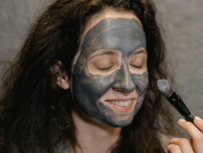 Mulher aplicando máscara de tratamento no rosto. As primeiras rugas surgem ao redor dos olhos, e os cabelos, antes brilhantes e escuros, começam a exibir fios prateados que surgem de maneira indomável, como se tivessem vontade própria. Não é algo necessariamente ruim, mas um lembrete de que a vida está acontecendo. É como se o espelho, antes cúmplice de nossa juventude, agora fosse um parceiro de uma realidade menos idealizada