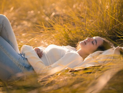 Uma mulher repousa sobre a relva iluminada pelo sol, em um momento de contemplação e bem-estar. O contato com a natureza remete ao convite de Gaia para reencontrar o pertencimento ao corpo vivo da Terra