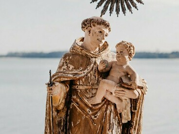 A representação de Santo Antônio ganha destaque diante da paisagem marítima, unindo a imagem religiosa ao ambiente natural. Na obra de Socorro Acioli, a figura do santo também ultrapassa o espaço da devoção e se torna parte do imaginário que dá vida à fictícia Candeia