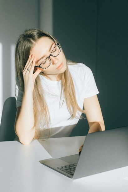 A young woman removes her glasses in a bright office after prolonged screen exposure, reflecting the fatigue and digital eye strain caused by constant device use