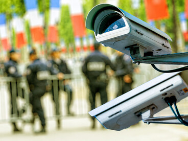 Police officers patrol beneath a mounted CCTV surveillance camera in a monitored public space with a blurred city background