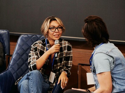 A woman sitting down with microphone in hand is speaking publicly in front of a group of eager listeners
