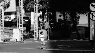 Sozinho, um menino atravessa a rua em direção à parte escura da imagem, deixando para trás a área iluminada onde seus passos ainda podem ser vistos. A cena evoca a trajetória de uma infância marcada não por uma perda, mas por uma ausência que já estava presente desde o nascimento com a ausência de um pai que nunca chegou a ocupar seu lugar