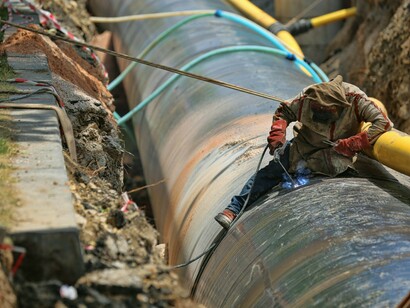 During the day in Turkey, a man in a black jacket and blue denim jeans is seated on a pipeline, working on repairs