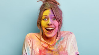 Headshot of an optimistic woman in a white t-shirt, smiling as she plays with vibrant colors, her face and clothes covered in colorful splashes