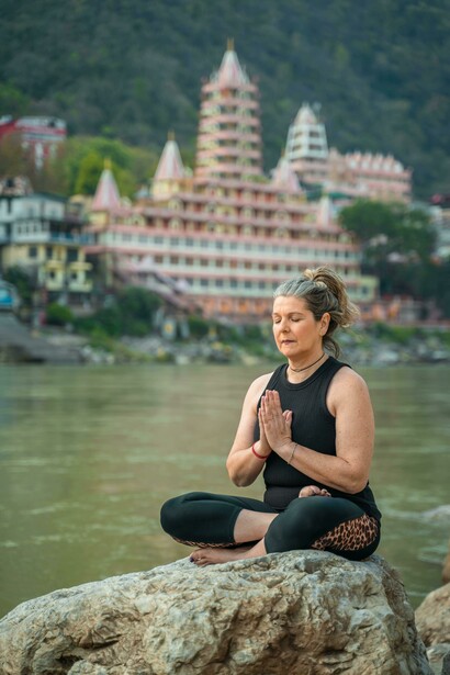 In Rishikesh, India, an elderly woman practices yoga on the riverbank, her eyes closed in deep meditation as the Ganges flows gently beside her