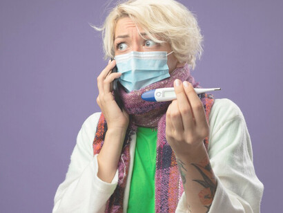 Unhealthy woman with short hair and a facial protective mask, holding a thermometer and talking on a mobile phone against a purple wall, symbolizing healthcare costs and economic challenges