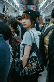 A young Korean woman stands on a subway train, gazing directly at the camera, surrounded by other commuters
