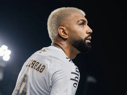 Em um enquadramento fechado, Gabigol aparece de perfil, com o olhar direcionado para cima e a camisa branca do Santos destacando o nome Gabriel e o número 9 nas costas. A iluminação do estádio contrasta com o fundo escuro e reforça a expressão concentrada do jogador. O retorno ao clube formador representa mais do que a volta a uma camisa conhecida: é a oportunidade de reencontrar o caminho das boas atuações e reafirmar seu valor no futebol brasileiro