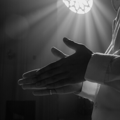 Unas manos masculinas en posición de rezo iluminadas por un haz de luz que entra desde el rosetón de una iglesia