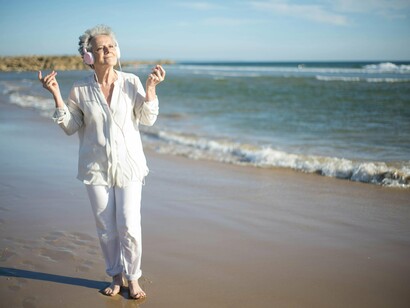 Mulher ouve música enquanto caminha na praia. Pense nas ondas delta como o BPM mais lento que existe. São essas ondas que dominam o nosso cérebro durante o sono profundo. Sabe aquele "after" quando você só quer relaxar e desligar? O som ambiente, como chuva ou sons suaves da natureza, ajuda a entrar nesse estado