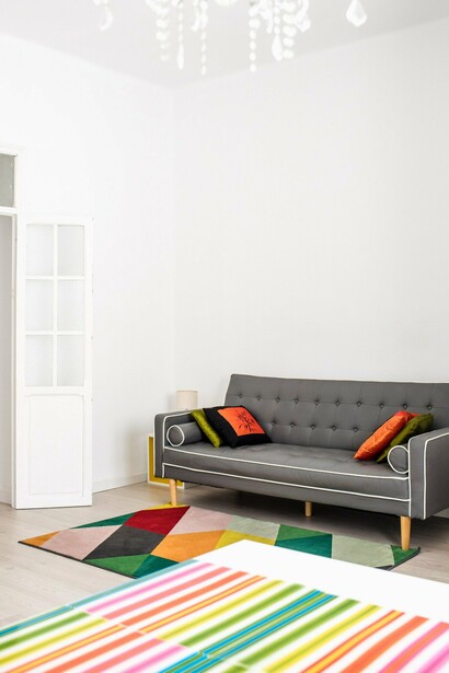 Uma sala ampla reúne sofá cinza, almofadas coloridas, tapetes de desenhos geométricos e um lustre de estilo clássico. A mistura de elementos evidencia como uma casa pode reunir diferentes referências e, ao mesmo tempo, tornar-se uma expressão da identidade de quem a habita