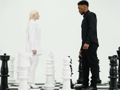 Two individuals stand face-to-face on a life-sized chessboard, their contrasting black and white outfits mirroring the chess pieces around them.