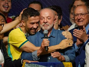 Durante o pronunciamento após a vitória, Lula segura um microfone enquanto André Janones, vestido com a camisa amarela da seleção brasileira e responsável pela criação do grupo de WhatsApp "Exército do Lula" que reunião quase 11 milhões de pessoas, o abraça. Ao redor, aliados acompanham o momento com expressões de alegria e gestos de apoio; diante deles, parte de uma bandeira nacional aparece junto à tribuna. A cena registra a proximidade física e emocional entre o presidente e os integrantes de seu grupo político. Entretanto, a vitória não elimina as divisões que atravessam a sociedade brasileira. Para Boaventura de Sousa Santos, o desafio que se abre é transformar o resultado das urnas em condições para reconstruir o diálogo democrático e enfrentar as forças que procuram restringir o pluralismo político