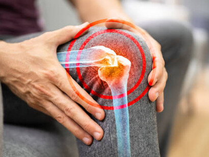 An elderly woman sits in her living room, holding her knee as she experiences arthritis and joint discomfort