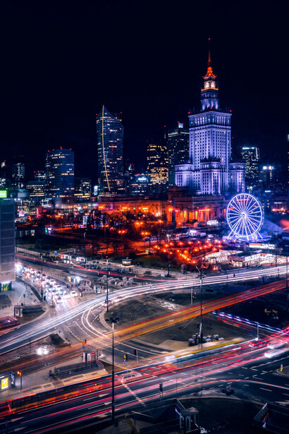 A nighttime view of Warsaw, illuminated by the glow of a Ferris wheel in the distance, highlighting the city's vibrant streets and dynamic urban energy