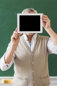 Un professore tiene un tablet con lo schermo nero nascondendo il viso, sullo sfondo una grande lavagna verde 