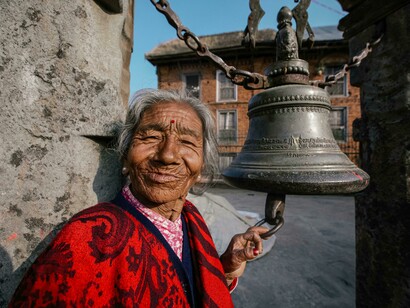 A smiling senior Indian woman stands beside an ancient temple bell in Bhaktapur, Nepal